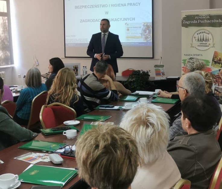 Inspektor pracy przemawia w sali szkoleniowej przed osobami siedzącymi przy stołach