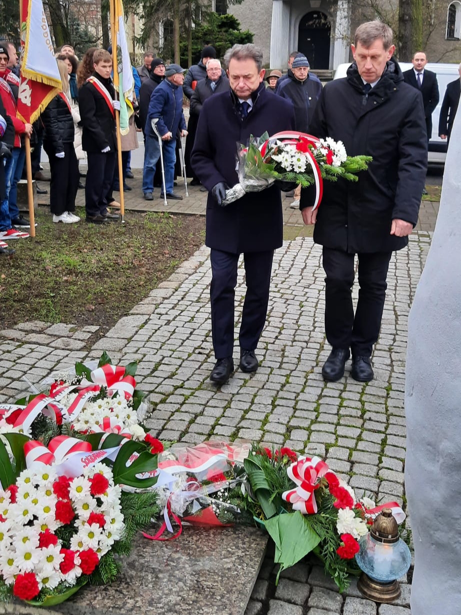 Jerzy Łaboński i Robert Simonjetz stoją na tle uczestników wydarzenia. Jerzy Łaboński trzyma wiązankę kwiatów