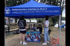 Widok na stoisko OIP. Niebieski namiot wystawienniczy, pod zadaszeniem stolik, ulotki, plakaty oraz osoby obsługujące i zainteresowani 