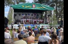 Na scenie stoją ludzie ubrani w stroje ludowe. Przed sceną siedzą uczestnicy imprezy