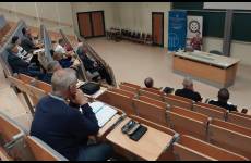 W sali wykładowej prelegent stoi przed uczestnikami spotkania siedzącymi w ławkach