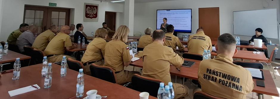 Widok sali szkoleniowej. Uczestnicy szkolenia siedzą przy stołach ustawionych w rzędach. Przed nimi siedzi omawiająca zagadnienie inspektor pracy. Za inspektor na ekranie jest wyświetlony slajd z prezentacji multimedialnej