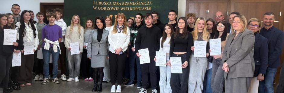 Zdjęcie uczestników konkursu z komisją konkursową. Wszyscy stoją na tle ścianki wystawienniczej Izby